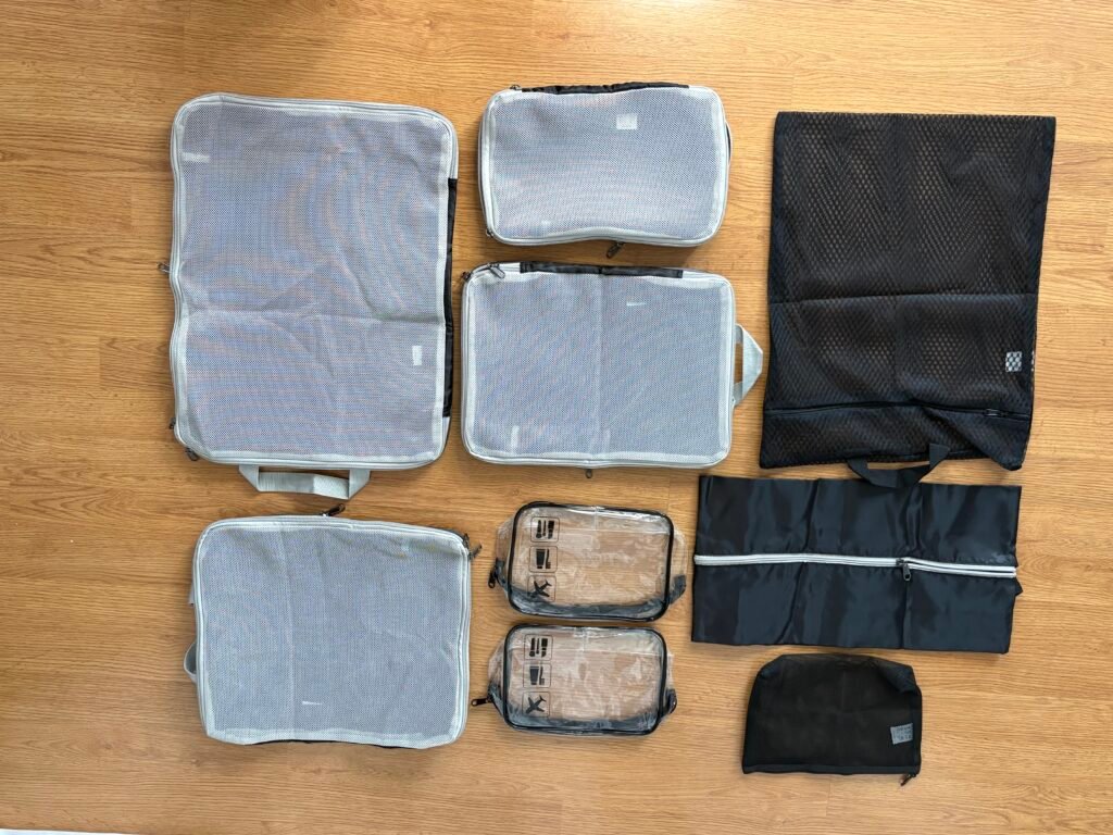 A collection of grey mesh packing cubes, clear toiletry bags, and black laundry pouches laid out on a light wood floor.