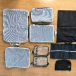 A collection of grey mesh packing cubes, clear toiletry bags, and black laundry pouches laid out on a light wood floor.