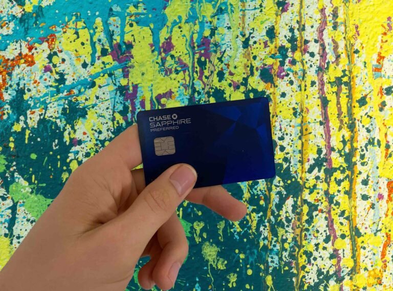 A hand holding a blue Chase Sapphire Preferred credit card against a vibrant yellow and teal painted background.