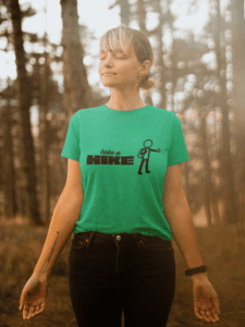 This lifestyle image features our “Take a Hike” graphic t-shirt, styled in a peaceful woodland setting that reflects the spirit of adventure and intentional time outdoors. The front design showcases a minimalist stick-figure hiker and bold lettering, while the back delivers a clever surprise message: “I Will Join You.” Made for hikers, campers, and nature lovers who appreciate humor with heart, this shirt is perfect for trail days, national park trips, casual weekends, or gifting to your favorite adventure buddy. Comfortable, versatile, and conversation-starting, it’s a wearable reminder that the best journeys are meant to be shared.