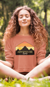 This image features the “Adventure Awaits” graphic sweatshirt, styled in a tranquil woodland setting that reflects mindful exploration and a love for the outdoors. The design showcases a warm sunset layered behind a mountain silhouette, framed in a clean geometric shape with the phrase Adventure Awaits—inviting both movement and stillness. Perfect for hikers, travelers, yogis, and nature lovers, this sweatshirt is made for slow mornings, forest walks, campfire evenings, and moments of quiet reflection. Soft, relaxed, and effortlessly wearable, it pairs adventure-inspired style with everyday comfort—reminding you that the journey begins wherever you are.