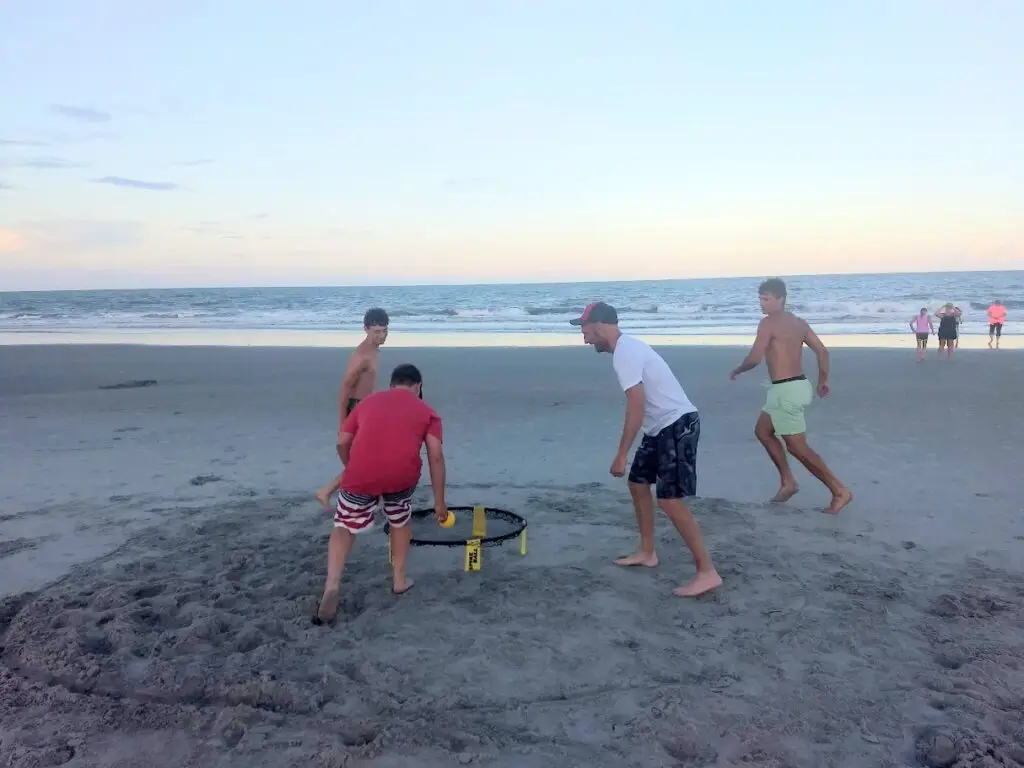 Four young men playing a fast-paced game of Spikeball on a wide, uncrowded beach in Murrells Inlet, South Carolina, with the ocean in the background at dusk.
