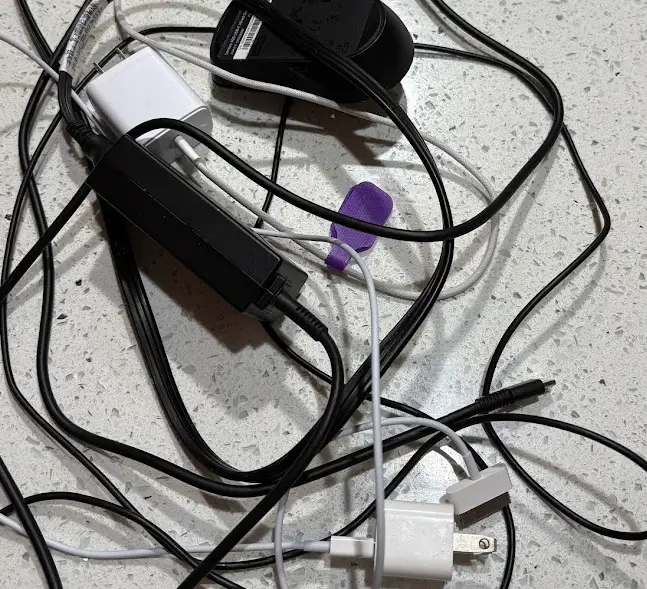 A cluttered pile of tangled white and black charging cords, power adapters, and USB cables on a wooden desk surface, illustrating the need for a charging cord organizer.