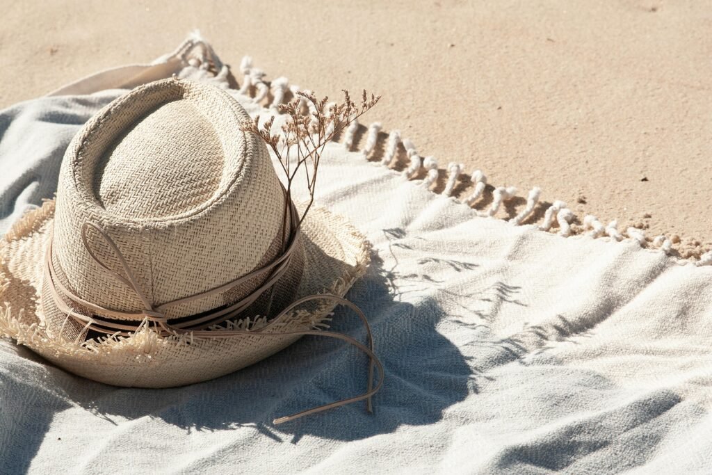 straw-hat-symbolizing-travel-memories-and-fashion. Close-up of a straw hat with plants on a beach blanket under bright summer sunlight. Memories and Fashion meet.