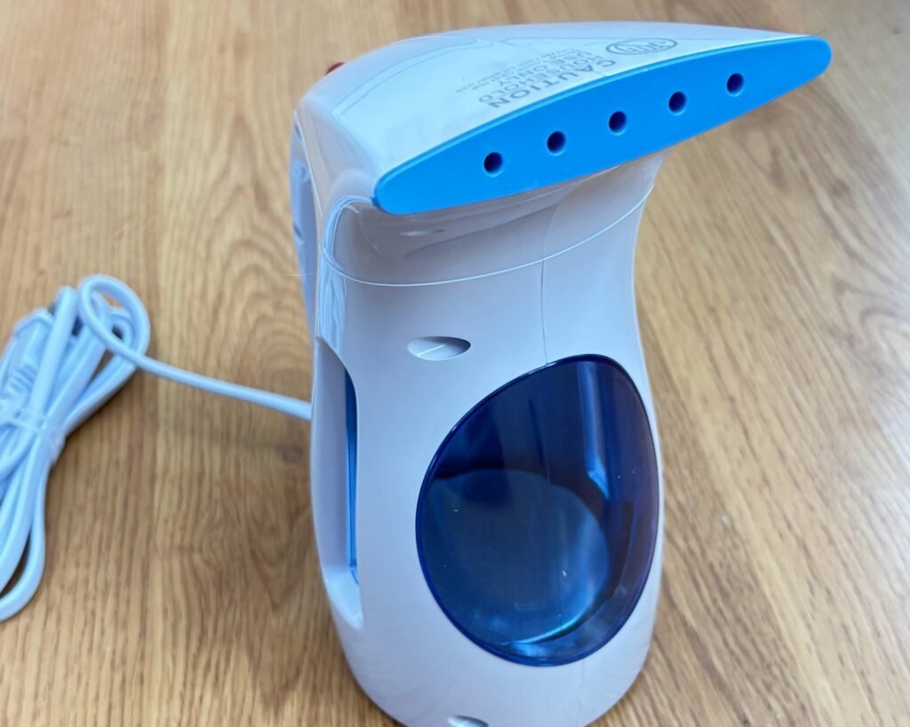 White and blue HiLife portable handheld garment steamer with a clear water tank on a wood floor.