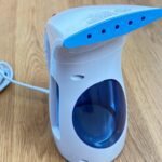 White and blue HiLife portable handheld garment steamer with a clear water tank on a wood floor.