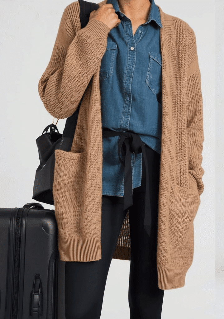 A woman wearing a comfortable airport outfit women need, including a long camel cardigan, denim shirt, leggings, and a carry-on suitcase for a stress-free trip.