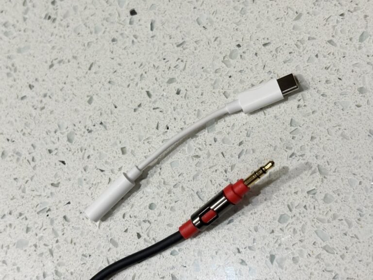 A white USB-C to 3.5mm audio headphone adapter next to a black and red audio jack cable on a white speckled surface. How to extend the life of beats.