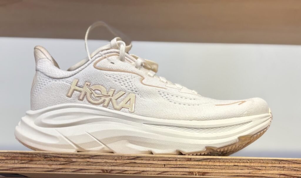 A side profile of a white Hoka Clifton 10 running shoe with gold accents sitting on a wooden shelf, described as one of the best travel shoes for carry-on travel.