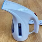 A blue and white HiLIFE portable handheld clothing steamer with a white power cord, sitting on a wooden floor.