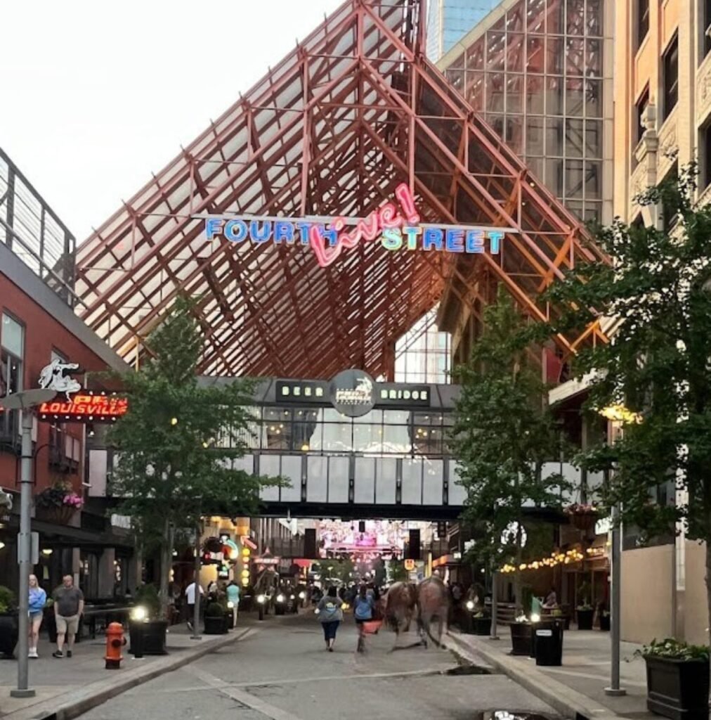 A pedestrian-only street scene at Fourth Street Live! in downtown Louisville, featuring a large red-framed glass canopy, outdoor dining patios, and a "Fourth Street" neon sign.
