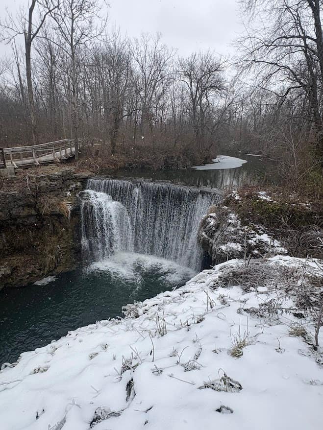 A high-angle view of Cedar Cliff Falls in Cedarville, Ohio, showing water flowing over a wide stone dam into a snowy riverbed with a wooden bridge in the background.