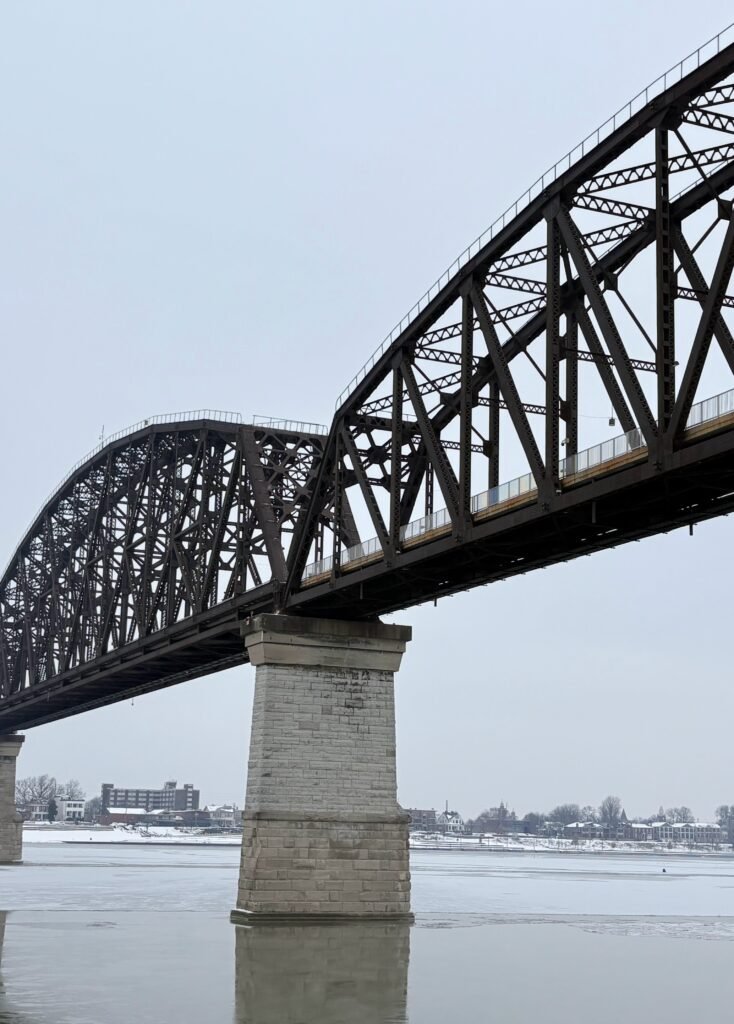 A snowy winter view of the Ohio River in Louisville, featuring the cable-stayed Abraham Lincoln Bridge, the Kennedy Memorial Bridge, and the Big Four Bridge truss in the far background under a gray sky.