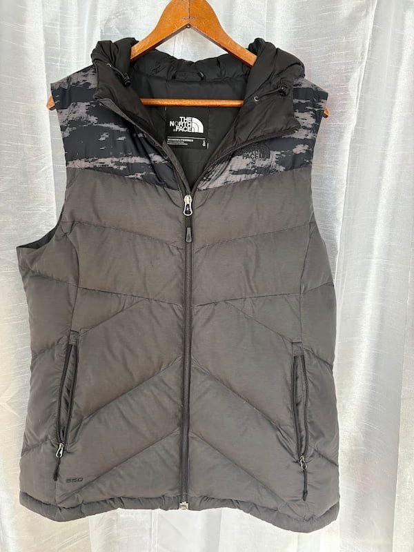 A dark grey and black quilted The North Face 550 down puffer vest with a hood, hanging on a wooden hanger as a windproof and water-resistant outer layer.