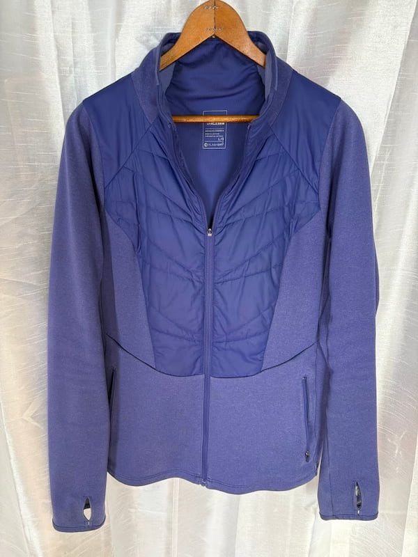 A purple hybrid jacket with a quilted, insulated front panel and stretch-knit sleeves, shown hanging on a wooden hanger as a mid-layer for winter hiking.