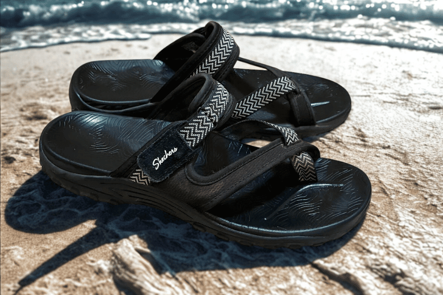 A pair of black Skechers Women's Reggae sandals with an adjustable back strap, sitting on a sandy beach, highlighting them as a versatile option for best travel sandals for women.