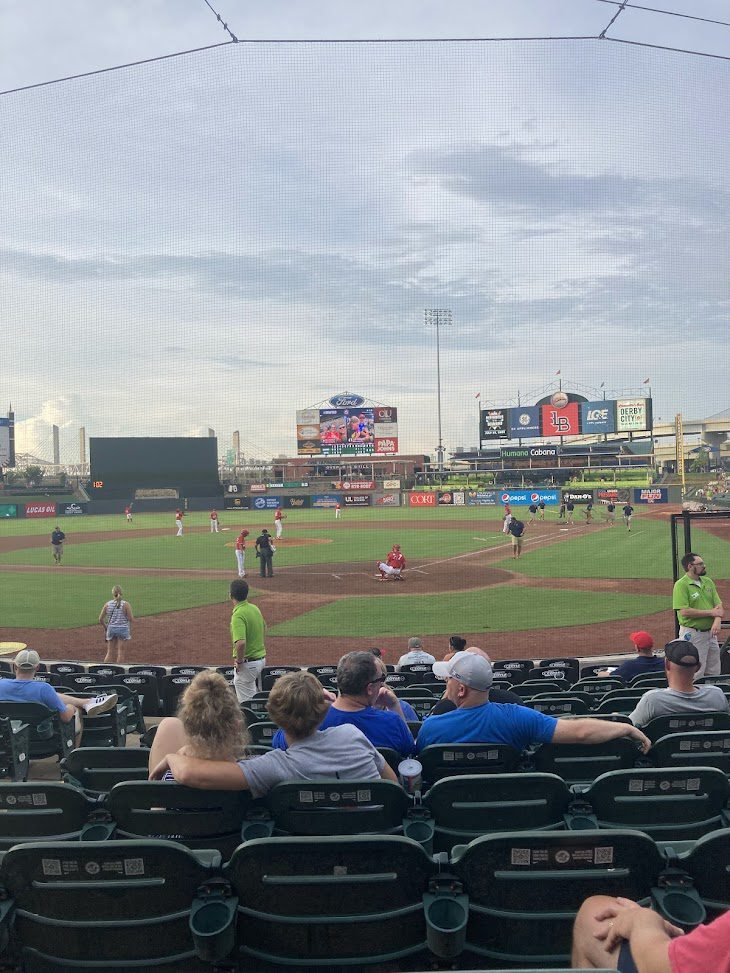 Interior view from the seating bowl of Louisville Slugger Field during a baseball game, showing the diamond, players, and outfield scoreboards under a cloudy sky.