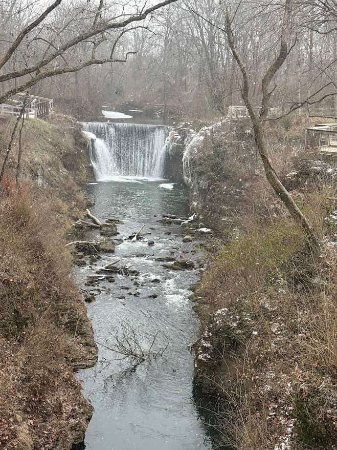 A high-angle view of Cedar Cliff Falls in Cedarville, Ohio, showing water flowing over a wide stone dam into a snowy riverbed with a wooden bridge in the background.