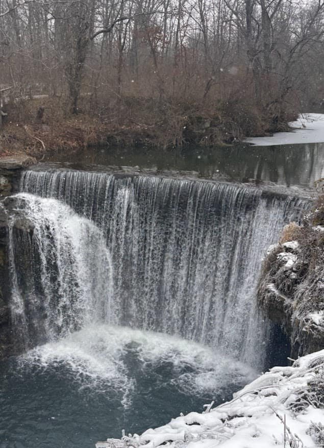 A close-up view of water rushing over the historic 1887 stone dam at Cedar Cliff Falls in Cedarville, Ohio, with mist rising from the river below.