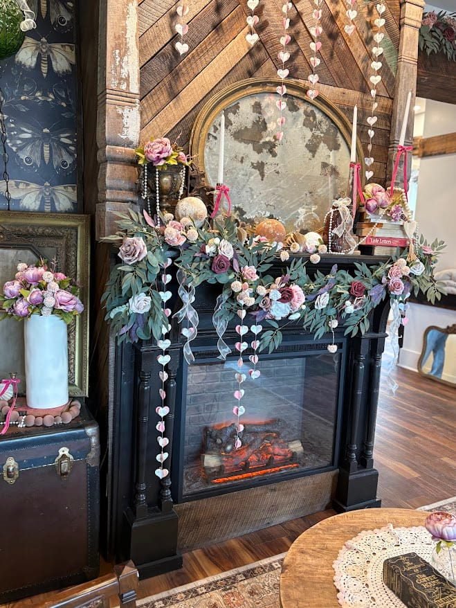 A cozy, romantic fireplace display at The Coffee House of Plymouth. The black mantel is draped with a lush garland of eucalyptus and roses in shades of dusty pink, white, and burgundy. Strands of small white and pink paper hearts hang vertically in front of the flickering electric fireplace. On the mantel, two tall white taper candles with red velvet bows flank a large, ornate gold-framed mirror that reflects the rustic wood-paneled wall behind it.