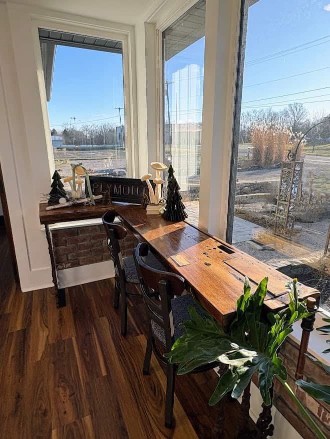 A bright window-side seating area in a coffee shop featuring a long, dark-stained wooden bar table. Two dark wood chairs with upholstered seats sit tucked under the bar, which overlooks a large window with a view of a street and a metal garden trellis. Festive holiday decorations, including small dark green Christmas trees, wooden mushroom figurines, and a sign that reads "PLYMOUTH," are arranged on the bar.
