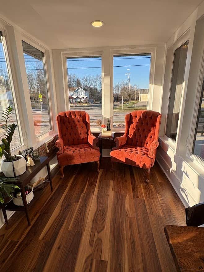 A bright, sunlit corner of The Coffee House of Plymouth featuring two large, vintage-style orange tufted wingback chairs. The chairs face each other on polished wide-plank walnut hardwood floors. Large windows surround the space, offering a view of the street outside and filling the room with natural light.