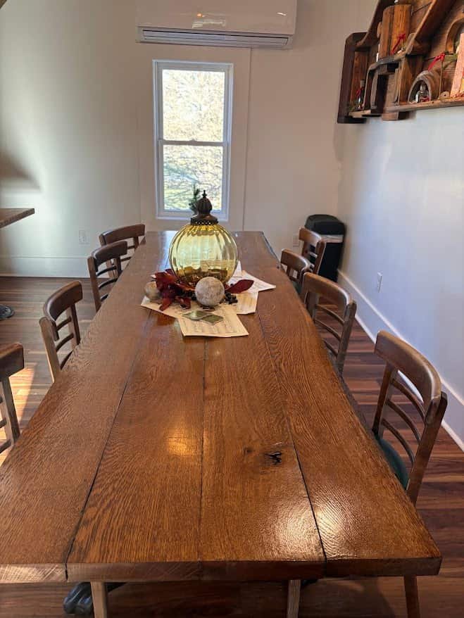 A long, rustic handcrafted wooden farm table made of wide oak planks, surrounded by matching wooden chairs in a bright room. The table is topped with a festive centerpiece featuring a large amber glass pumpkin, sheet music, and a white decorative orb. In the background, a shelf with rustic holiday decor hangs on the wall next to a large window.