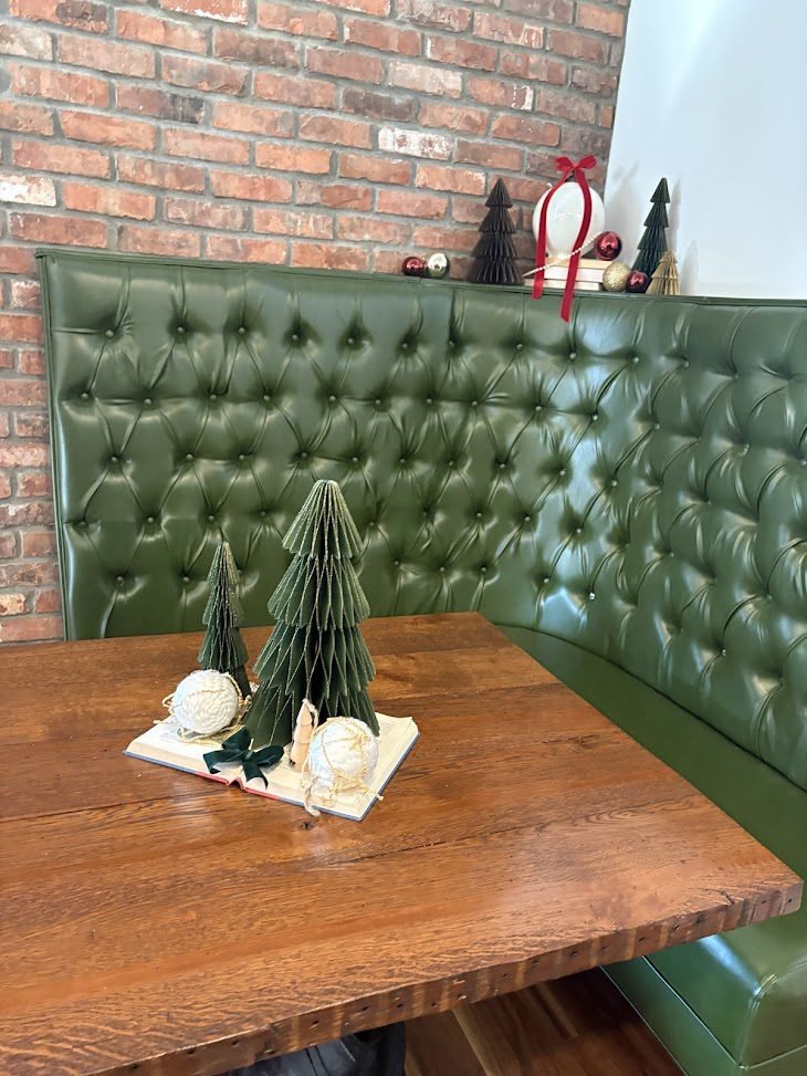 A cozy restaurant booth at The Coffee House of Plymouth featuring a deep green, diamond-tufted leather backrest against a red brick wall. On a rustic wooden table, two green paper accordion-style Christmas trees are arranged on an open vintage book, accented with cream-colored twine baubles and a dark green velvet ribbon.