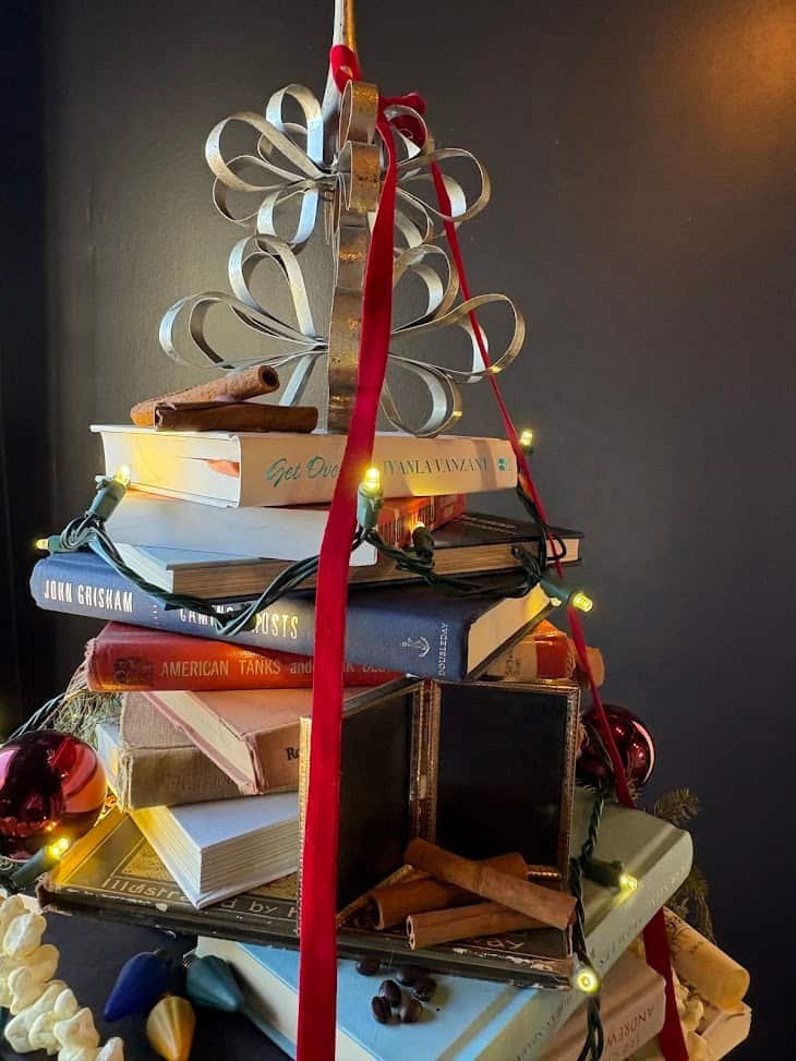 A "Christmas tree" made from a stack of vintage hardcover books, decorated with a strand of warm yellow fairy lights, red velvet ribbons, cinnamon sticks, and popcorn. A small, open decorative case and red Christmas ornaments are nestled within the stack, which is topped with a tiered, scrolled silver metal ornament.