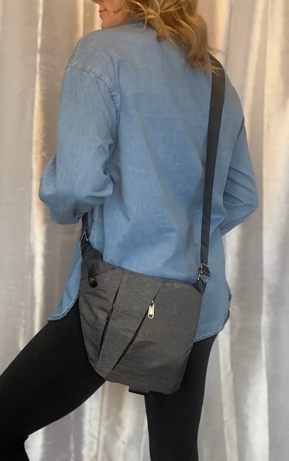 A woman wearing the best anti theft crossbody bag for travel in charcoal gray over a light blue denim shirt, showcasing its slim, ergonomic design.