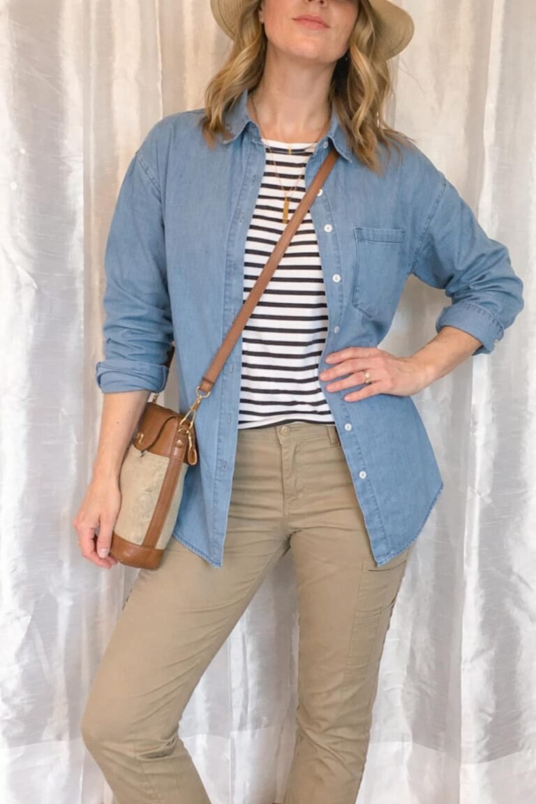 A woman modeling a stylish travel outfit featuring the best denim button down for travel worn open over a striped tee with tan cargo pants and a tan sun hat.