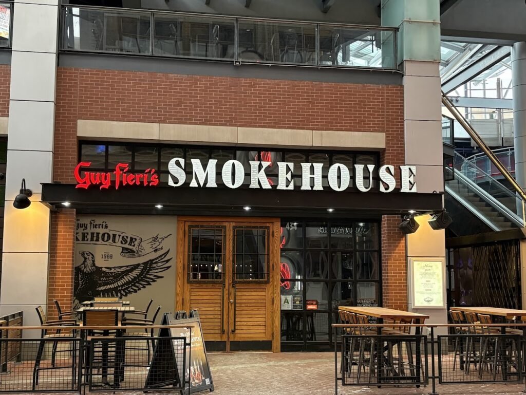 Exterior of Guy Fieri's Smokehouse at Fourth Street Live! in downtown Louisville, featuring a brick facade, large "SMOKEHOUSE" sign, and an outdoor patio with wooden tables.