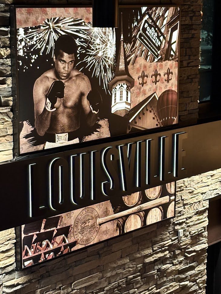 A vibrant, mixed-media wall collage in Louisville, Kentucky, featuring a black-and-white photo of boxer Muhammad Ali, a steeple from Churchill Downs, and illustrations of bourbon barrels and the Old Forester distillery logo.Louisville Travel Guide is here