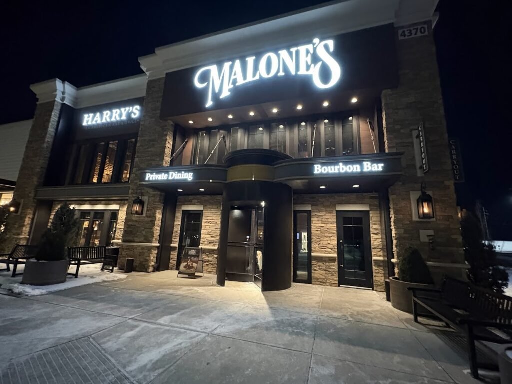 Nighttime exterior of Malone’s Prime Steakhouse and Harry’s American Bar & Grill at the Paddock Shops in Louisville, featuring illuminated signs, a stone facade, and a modern revolving door entrance.