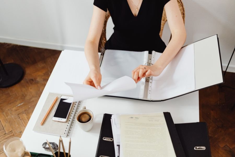 A person organizing documents into a black ring-binder physical travel document organizer with a coffee and smartphone on the desk.