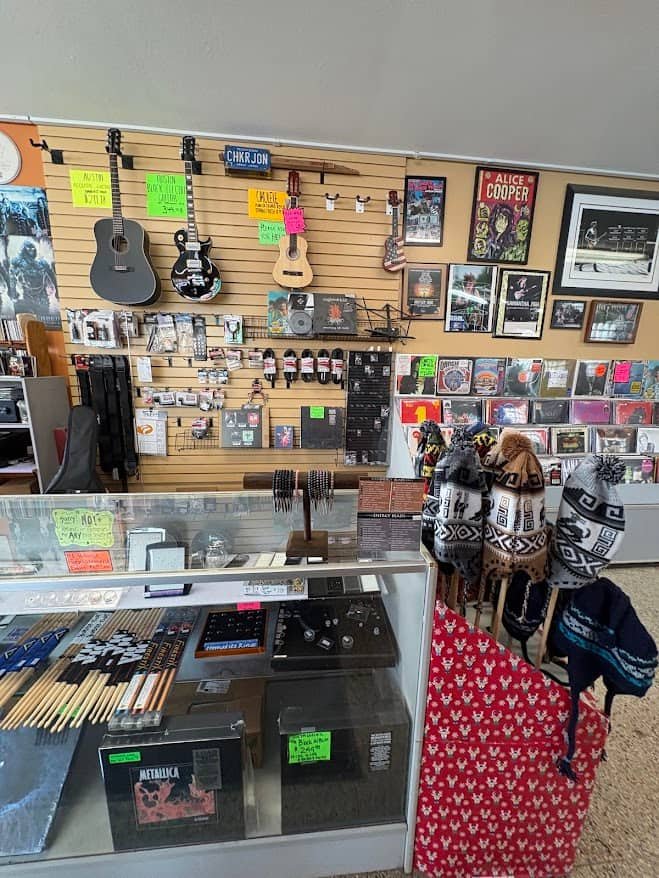 A retail display at Checker Records in Hillsdale, MI, featuring acoustic and electric guitars on a slatwall, a glass counter with accessories, and a rack of knit winter hats.