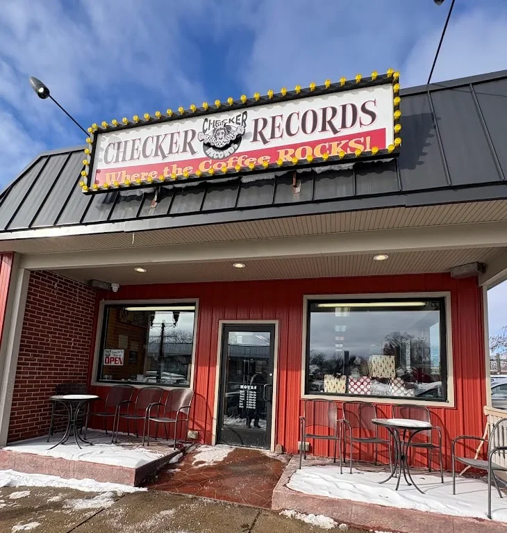 The vibrant red storefront of Checker Records in Hillsdale, Michigan, a relaxing coffee shop and record store featuring a "Where the Coffee Rocks!" sign and outdoor seating with a dusting of snow.