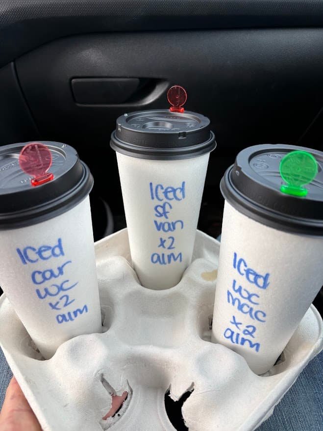 Three iced coffees with almond milk in a cardboard carrier inside a car, labeled with blue marker for a Midwest road trip.