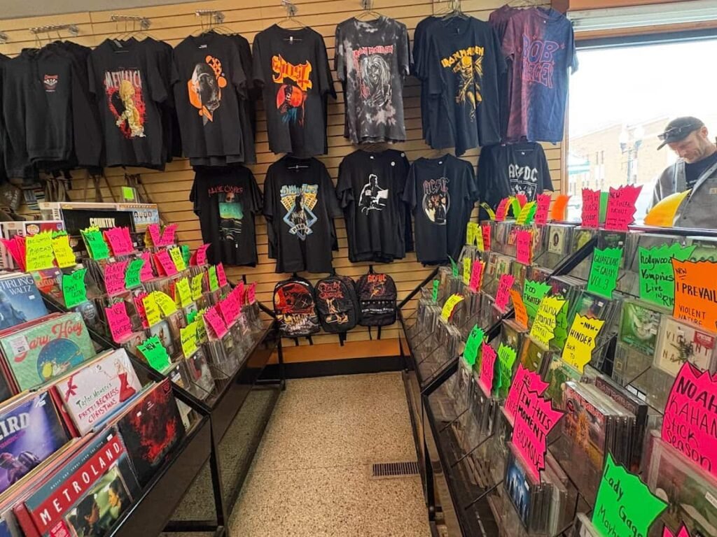 A vibrant display at Checker Records in Hillsdale, Michigan, featuring black band t-shirts on a slatwall and bins of CDs and vinyl with bright neon price tags.