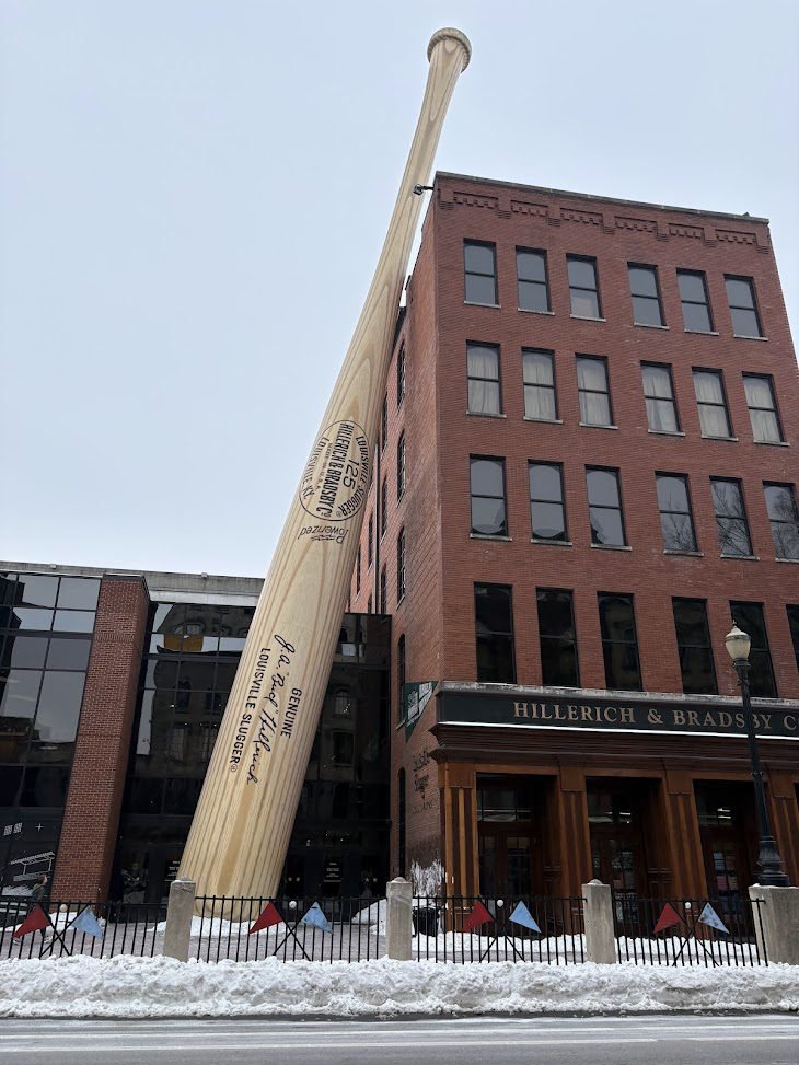 The world’s largest baseball bat, a 120-foot steel sculpture, leans against the red brick Louisville Slugger Museum and Factory in downtown Louisville under a cloudy sky with snow on the ground.