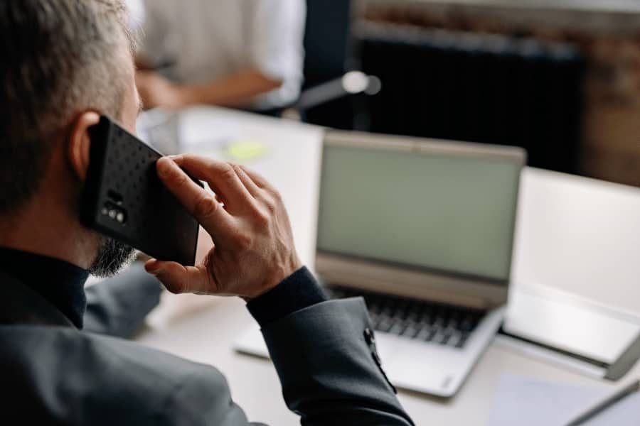 A man in a grey sweater sits at a wooden desk in a home office, talking on his smartphone while reviewing flight change documents for a Hawaii trip.