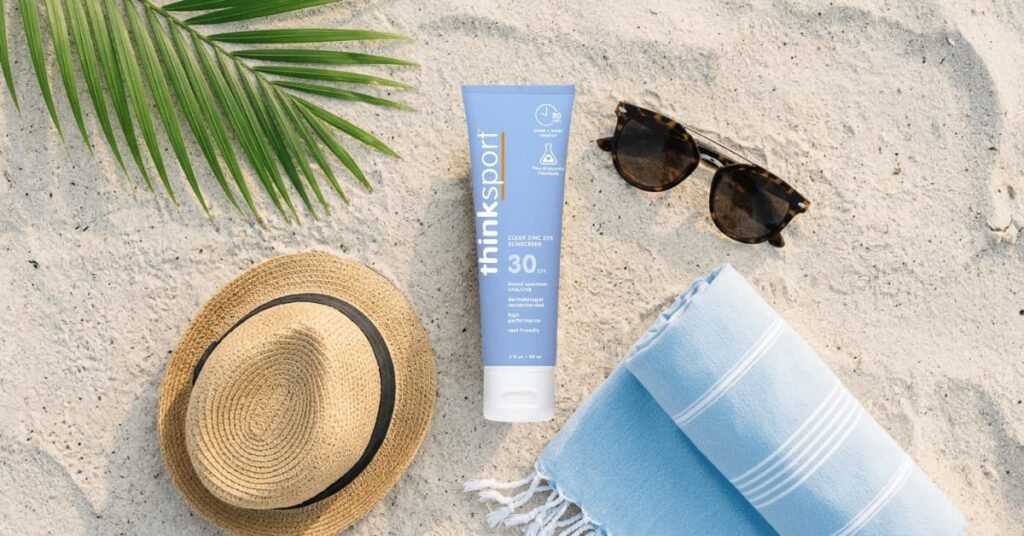 A flat lay on a sandy beach featuring a tube of Thinksport SPF 30 Clear Zinc mineral sunscreen, the best reef-safe sunscreen for beach travel, alongside tortoise-shell sunglasses, a light blue Turkish towel, and a straw hat.