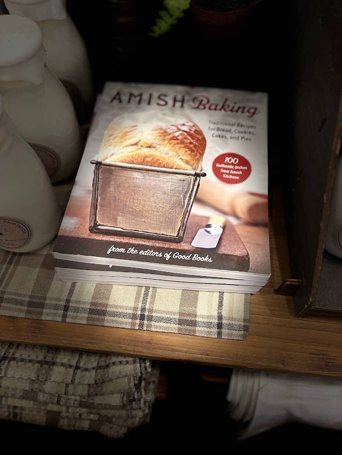 A stack of "Amish Baking" cookbooks on a wooden shelf in a store in Berlin, Ohio. The cover shows a fresh loaf of bread in a tin and notes "100 Authentic Dishes from Amish Kitchens."