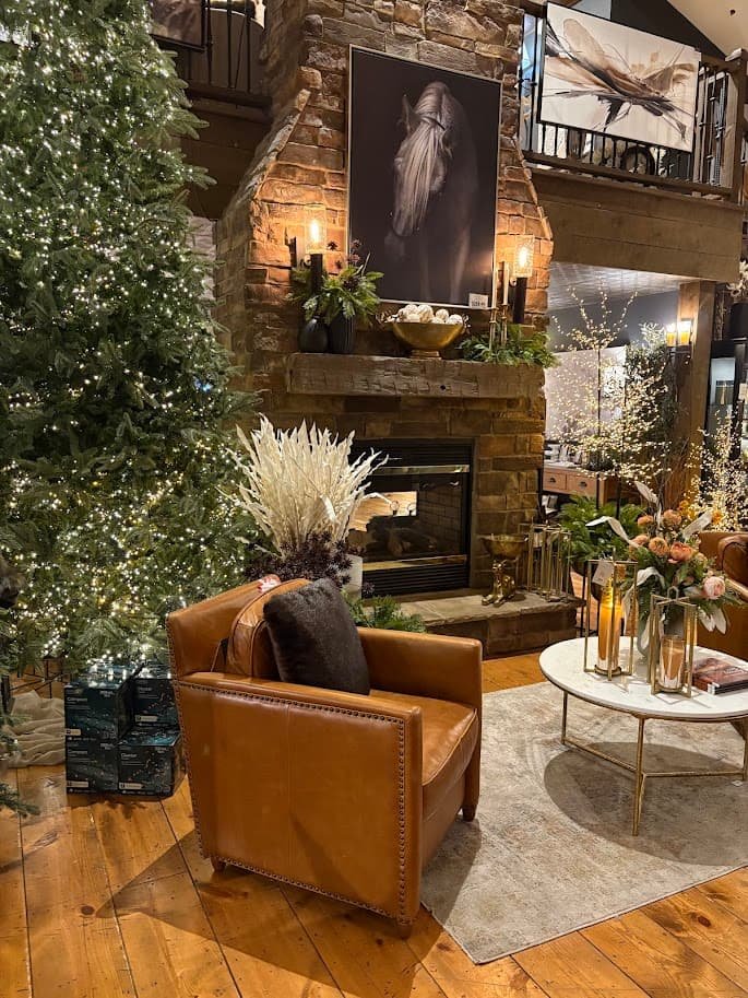 Cozy living room display inside the Village Gift Barn in Berlin, Ohio. Features a large stone fireplace, a brown leather armchair with gold studs, a lit Christmas tree, and a horse portrait hanging above the mantel. A perfect Day trip to Amish country.