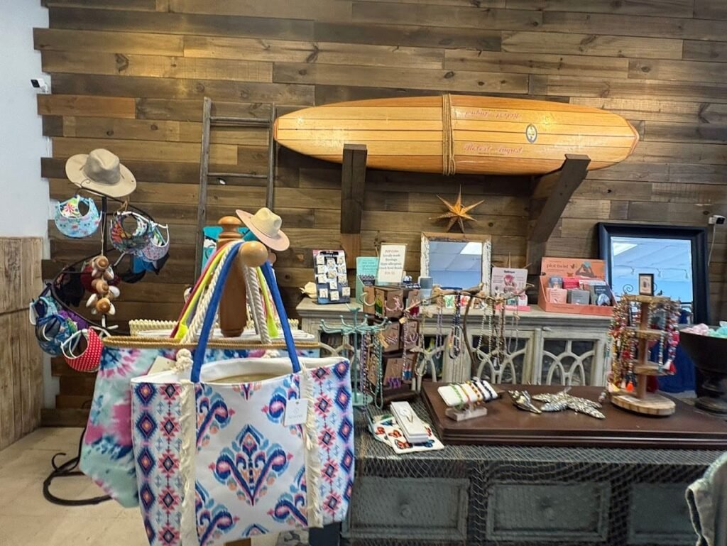The interior of Charmed By The Sea, a boutique on Flagler Avenue in New Smyrna Beach. Brightly patterned tote bags and beach hats are displayed on a table in front of a rustic wooden wall with a surfboard and mirrors.