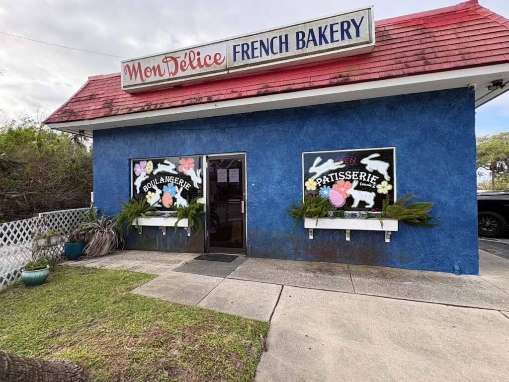 The exterior of Mon Delice French Bakery in New Smyrna Beach, featuring a blue building with a red roof and windows decorated with "Boulangerie" and "Patisserie" signage and Easter-themed bunny art.