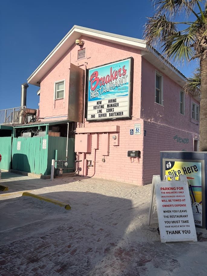 The exterior of The Breakers Restaurant, a bright pink two-story building with a large vintage-style neon sign, located right on the sandy shore of New Smyrna Beach under a clear blue sky.