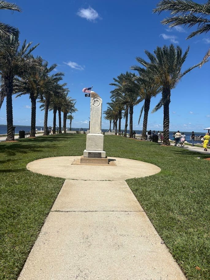 A tall stone war memorial monument at Riverside Park in New Smyrna Beach, Florida, flanked by rows of tall palm trees under a bright blue sky with a view of the Indian River in the background.