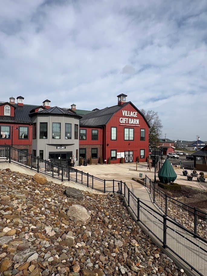 Exterior view of the Village Gift Barn in Berlin, Ohio. The large red and grey barn-style building features a stone walkway, black railings, and a large sign on the side.