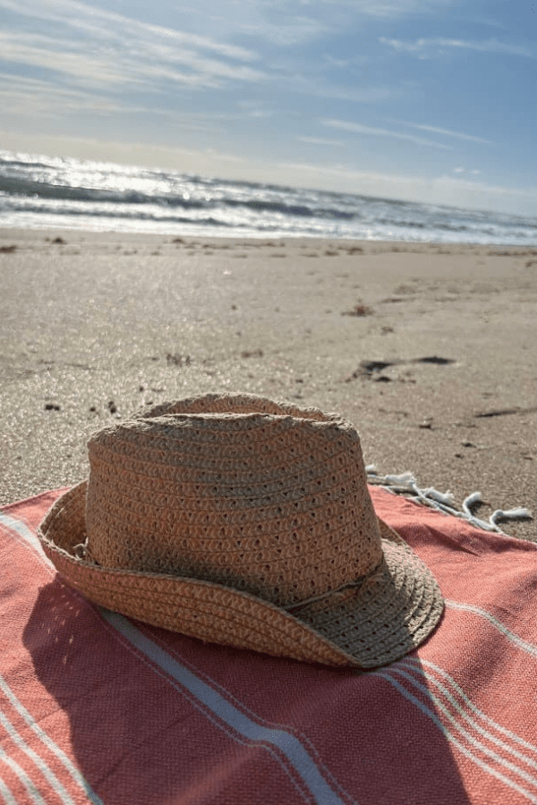 A straw sun hat resting on a pink and white striped BAY LAUREL Turkish beach towel on the sandy shore of New Smyrna Beach, with the sparkling ocean in the background.
